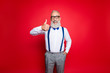 © deagreez - Portrait of cheerful, laughing old barber, stylist showing thumb up holding hand in pocket having blue suspenders, pants, bowtie, isolated on red background