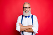 © deagreez - Portrait of professional, cool, old man with beaming smile having his arms crossed, looking at camera, wearing blue bow-tie and suspenders, isolated on red background