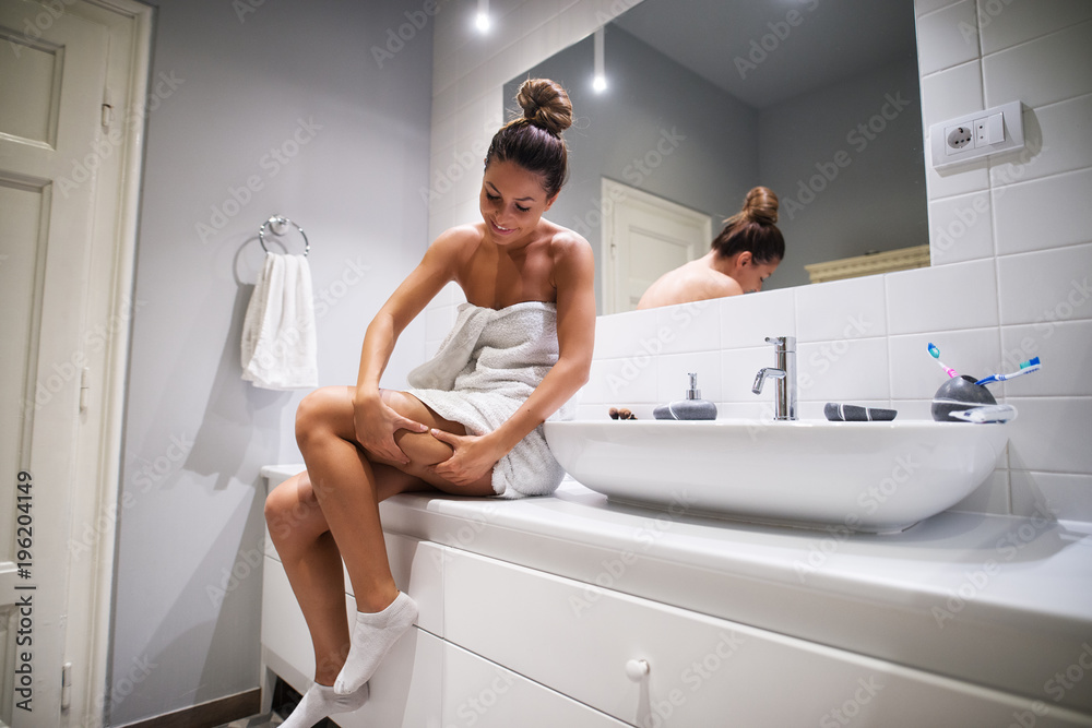 Young cute girl checking cellulite on the leg while sitting on bathroom elements in a towel before showering. - obrazy, fototapety, plakaty 