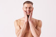 Portrait of young naked man applying aftershave and smiling while standing isolated on white background. Bearded hispter man standing shirtless and touching his face with hands. Men's skin care