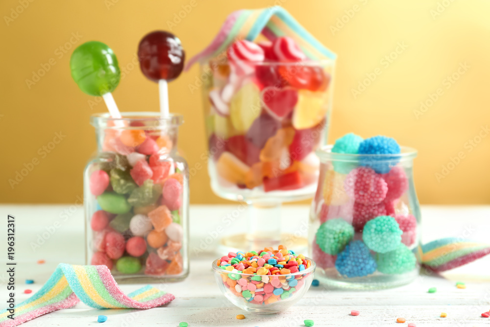 Glassware with sprinkles and different candies on table against color background