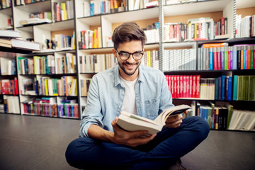 Front view of young hipster smiling man reading a book on the library floor.