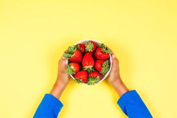 Poster - Kid holds in hand bowl with fresh strawberries on yellow background