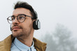 Close up portrait of handsome smart young Caucasian male wearing eyeglasses, looking at one side with headphones on misty nature background. Lifestyle, people and technology concept.