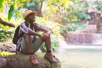 Wall Mural - African Man Traveler with backpack sitting and smiling with waterfall background.