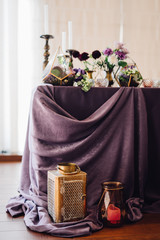 Wall Mural - Festive table decorated by violet cloth, flowers and greenery, candles on candleholders with golden lantern and candles near them. Wedding decor