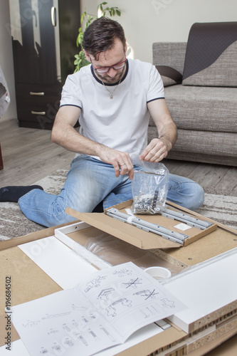 Fotografie, Tablou Mężczyzna klęczy na dywanie nad kartonem z elementami mebli do samodzielnego montażu i rozcina nożyczkami torebkę foliową z wkrętami i śrubami do montażu