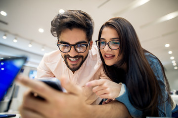 Wall Mural - Close up portrait view of adorable happy satisfied hipster young love couple holding and testing new mobile in a tech store.