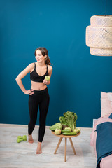 Wall Mural - Young woman exercising indoors with green healthy food on the table on the blue wall background at home