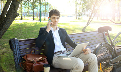 Businessman on a coffee break. He is sitting on a bench and working at laptop, next to bike.