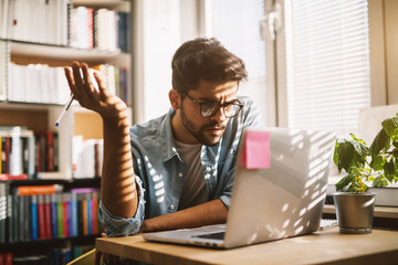 Wall Mural - Bearded nerdy university student having trouble while studying for an exam on a laptop in the library.