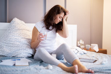 Tired Ill Middle Aged Woman Holding Hand On The Forehead While Having The Glass Of Water In Another Hand And Paper Wipes Are Around Her On The Bed.