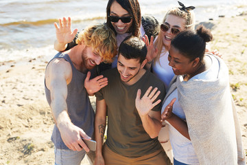 Wall Mural - Cheerful friends waving hands while recording video or making selfie in smartphone while enjoying summer day on the beach