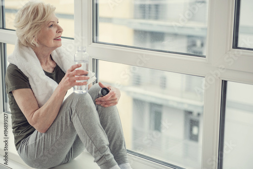 Fotografía Good looking old woman sitting on windowsill with bottle in hand, she is looking out the window with smile