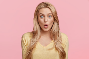 Wall Mural - Isolated shot of pretty young female stares at camera with shocked expression, recieves unexpected news, keeps lips rounded, poses in studio over pink blank wall. Great surprisment or unexpectedness