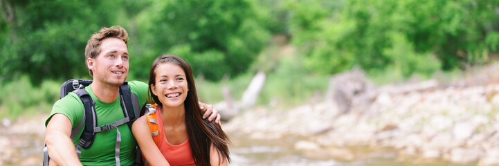 Wall Mural - Happy hikers couple hiking in nature forest outdoors smiling portrait of interracial young people with backpacks camping in summer outdoor. Banner panorama.