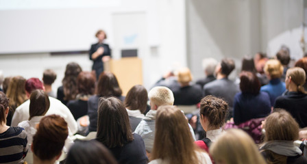 Female speaker giving presentation in lecture hall at university workshop. Audience in conference hall. Rear view of unrecognized participant in audience. Scientific conference event.