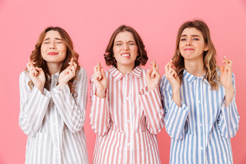 Sticker - Portrait of three joyful young girls 20s wearing colorful striped pajamas with closed eyes willing good luck keeping fingers crossed during sleepover, isolated over pink background