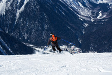 Snowboarder In A Mask And Gear Roll On The Board Down The Hill With His Back To The Camera. Below The Mountain Forest And The Snow-covered Valley