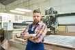 © Seventyfour - Waist up portrait of young factory worker looking at camera while standing with arms crossed posing by wood cutting machine in modern workshop, copy space