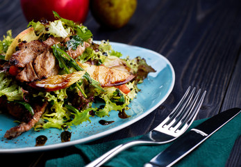 Veal salad with lettuce, apple, pear and tomato on blue plate on dark wooden background, copy space. Restaurant food, close up. Meat salad