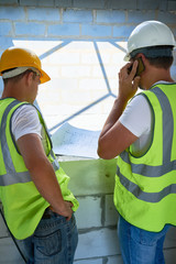 Wall Mural - Back view  portrait of two construction workers wearing hardhats discussing floor plans inside unfinished  building