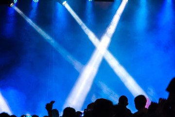 Concert crowd attending a concert, people silhouettes are visible, backlit by stage lights. Raised hands and smart phones are visible here and there.