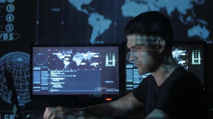 Male hacker programmer working at computer while blue code characters reflect on his face in cyber security center filled with display screens.
