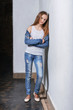 Fashion teenager girl standing leaning back against a white wall. Young caucasian woman model in casual stylish clothes, denim jacket, white singlet cotton, youth jeans. High fashion urban style.