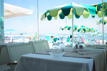 There is a mediterranean restaurant with tables covered with white tablecloths. Sun umbrellas in green tones and the sea beach are located in the background. The atmosphere is cool and serene.