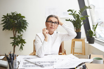 Portrait of pensive gray haired female architect in her fifties touching head while working at her office desk, making drawings using architectural tools, looking up, searching for inspiration