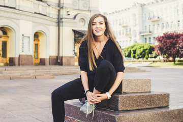 cute laughing model in black jumpsuit sitting at the street and trying to tie shoelaces