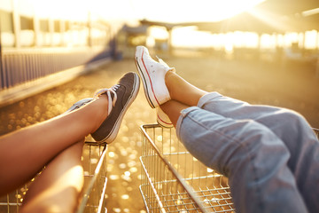 Nice legs! Two beautiful girls having fun on shopping trolleys. Multiethnic young people racing on shopping cart. Beautiful summer day with sunlight. 