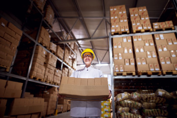 Young tensed female worker in sterile cloths and yellow helmet is carrying a very heavy stack of brown cardboard boxes from a facility storage room.