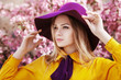 Outdoor close up portrait of young beautiful fashionable girl posing in street with blooming trees, wearing stylish wide-brimmed violet hat, yellow shirt, bow tie. Female spring fashion concept