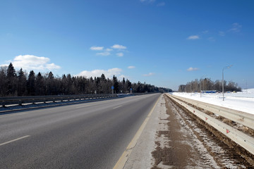 Landscape with long straight suburban highway with metal a fence on the sides and forest under clear blue sky on sunny winter day