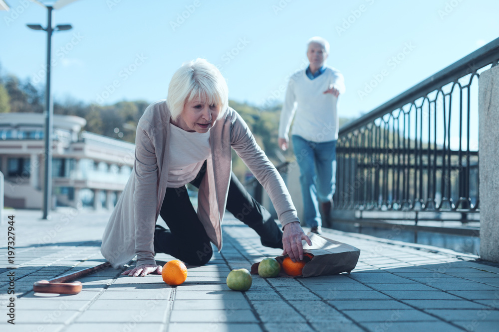 Are you ok. Selective focus on a scared senior lady standing on her knee and trying to pick up her groceries after falling down while her worried husband running to her in the background. - obrazy, fototapety, plakaty 