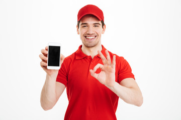 Wall Mural - Excited happy young delivery man showing display of mobile phone make okay gesture.