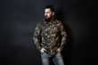 Fashion portrait of young bearded man in trendy military shirt and blue jeans over black backround. Stylish casual look.