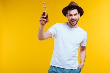 cheerful young man in hat holding glass bottle of summer drink and smiling at camera isolated on yellow