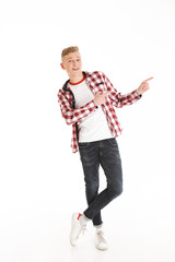 Poster - Full length portrait of happy educated teenage boy 17y wearing plaid shirt smiling and pointing fingers aside on copyspace, isolated over white background