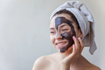 Wall Mural - Beauty portrait of a smiling brunette woman in a towel on the head applying black nourishing mask on face on white background isolated. Skincare cleansing spa relax cosmetics concept