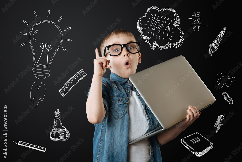 Great idea. Clever attentive creative child looking surprised and pointing his finger up while holding a big laptop and getting a brilliant idea - obrazy, fototapety, plakaty 