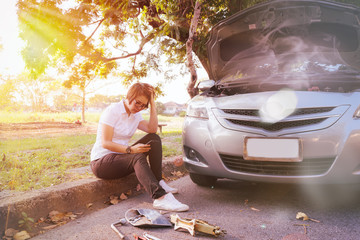 Woman sitting on ground and using mobile phone near her car broken down on the road side.