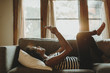 © Cavan Images - Side view of woman using smart phone while lying on sofa against window at home