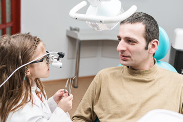 Canvas Print - Child in the role of a dentist in glasses-microscope treats an adult male seated in a dental chair