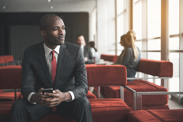 Waist up portrait of calm african guy sitting on couch and holding mobile phone in hands. Group of people resting on background. Copy space in right side