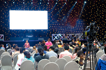 Professional camera recording on stage of business seminar meeting with projector screen and spotlights at the conference room, rear view of audience in the conference hall.