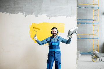 Wall Mural - Portrait of a happy repairman listening to the music with headphones during the painting process indoors