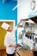 © pressmaster - Young woman in chef uniform taking frying pan from stack on top shelf before cooking